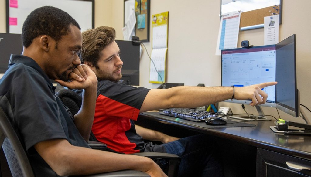 two men sitting in front of a computer , one of the men is pointing at the screen.