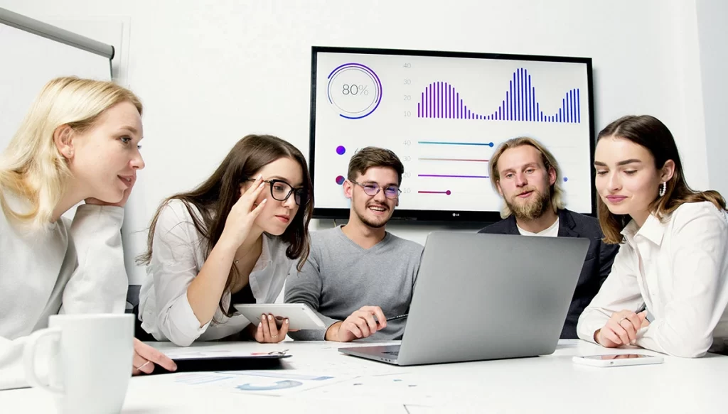 group sitting around a laptop which a chart with graphics is in the background